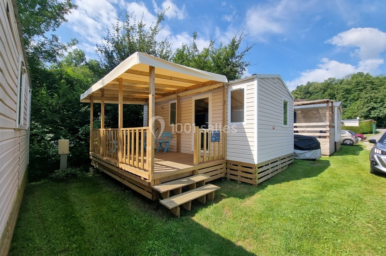 Mobil-home avec terrasse en bois, situé sur une pelouse entourée de végétation sous un ciel ensoleillé.