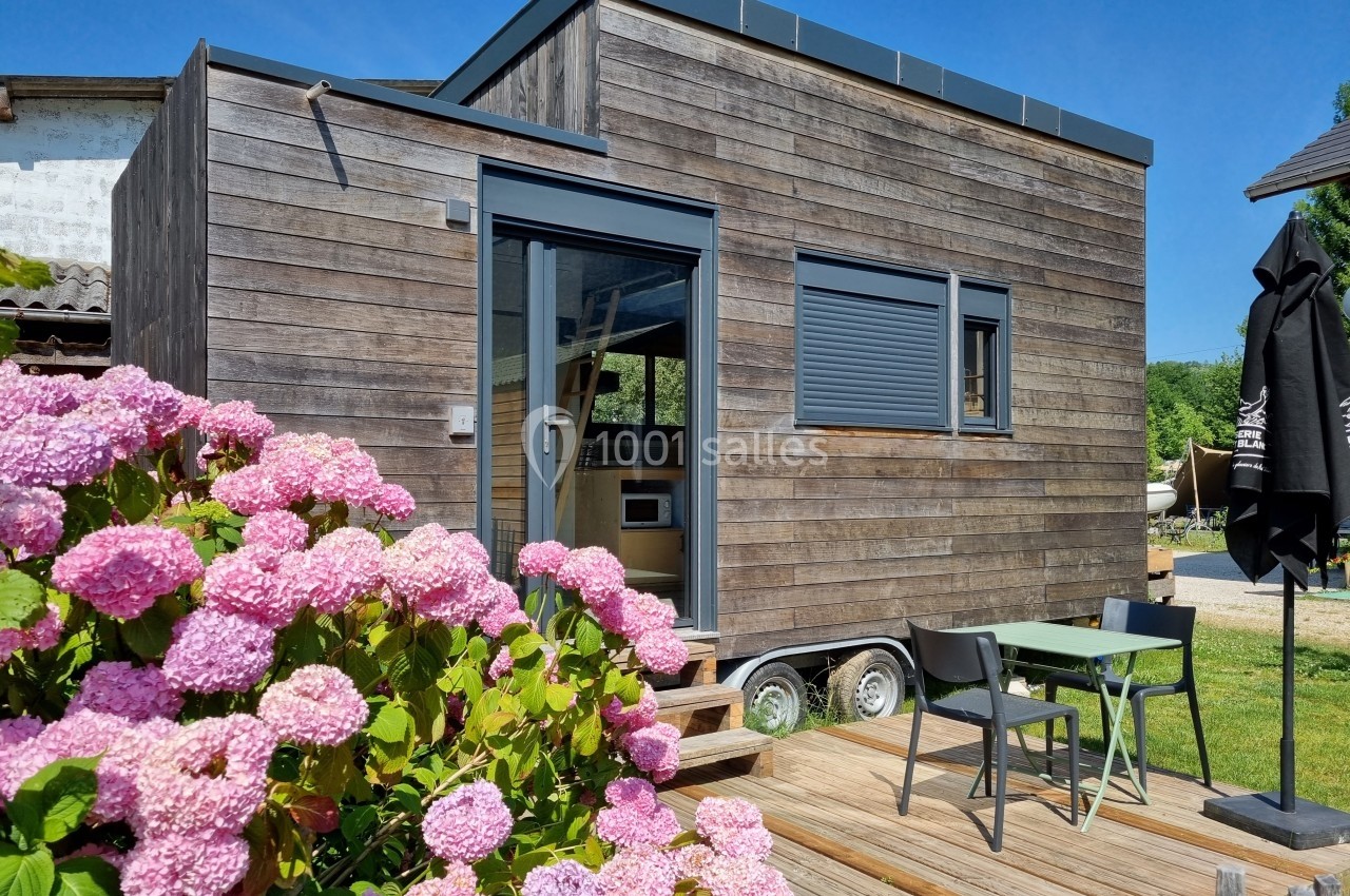 Petite maison en bois sur roues avec terrasse, table et chaises, entourée de fleurs roses et d'un paysage ensoleillé.