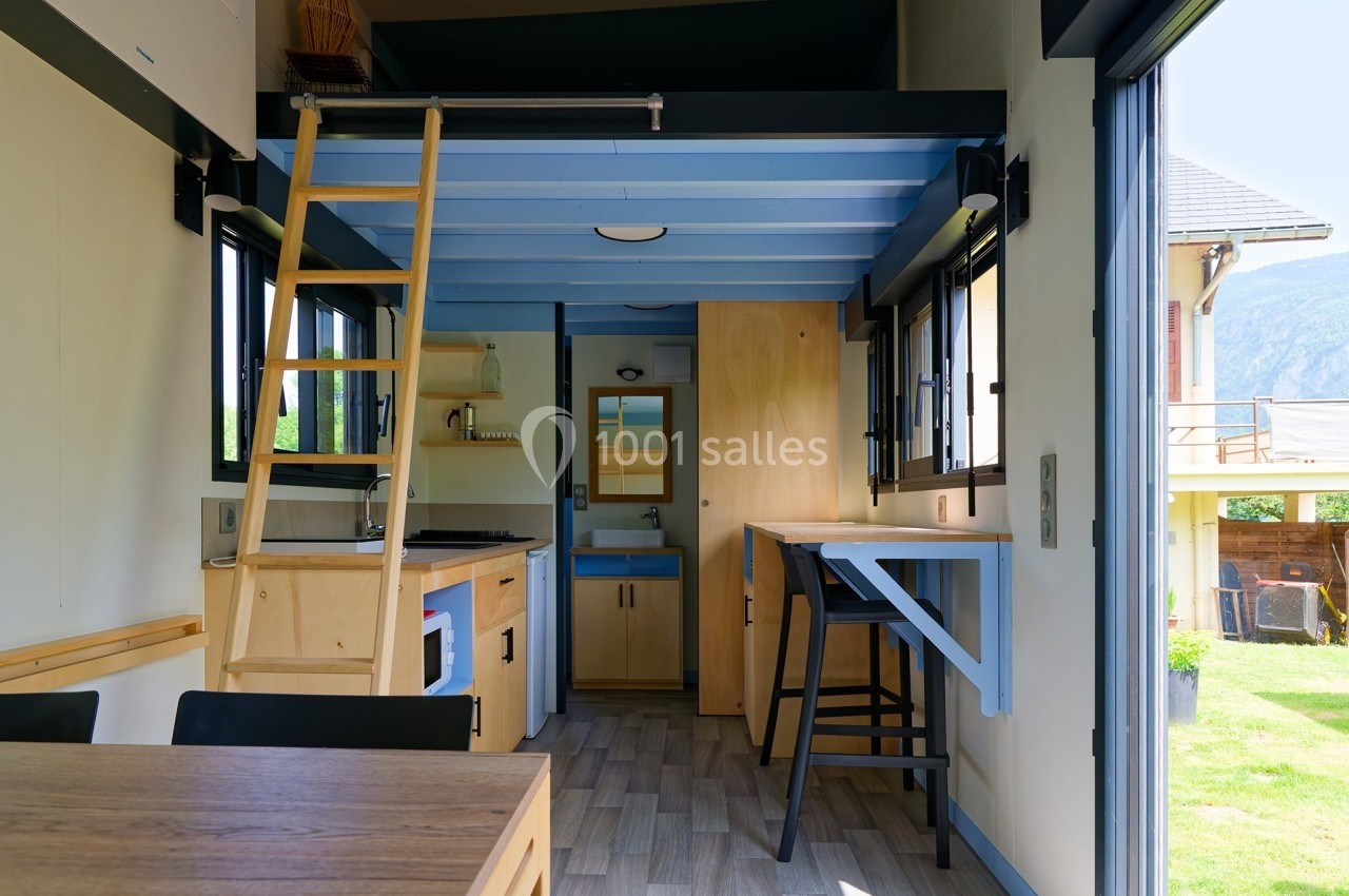 Intérieur d'une tiny house avec mezzanine, cuisine équipée, table pliante et vue sur l'extérieur par une baie vitrée.