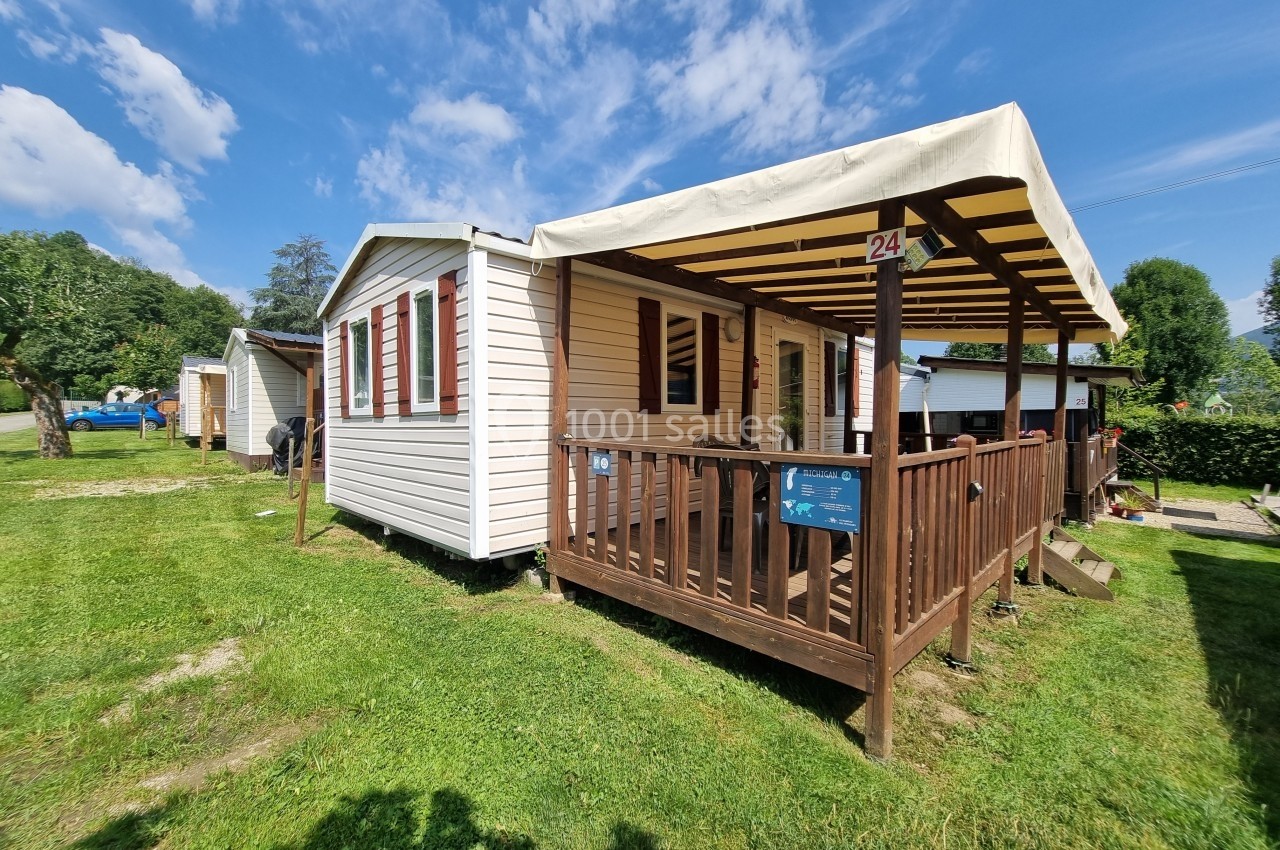 Mobil-home avec terrasse en bois et auvent, situé sur une pelouse verdoyante par une journée ensoleillée.