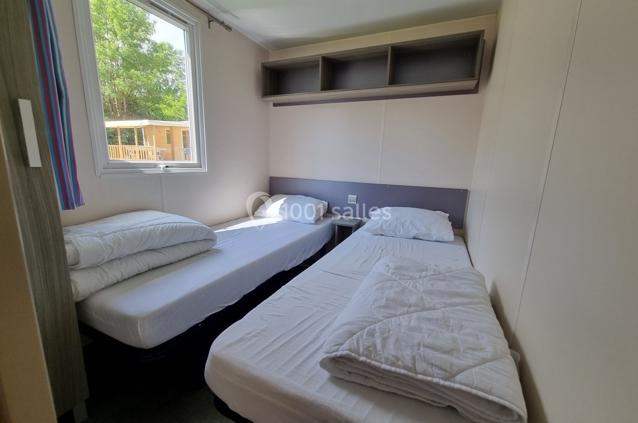 Deux lits simples avec draps blancs dans une chambre lumineuse, une fenêtre donnant sur l'extérieur et des étagères murales.