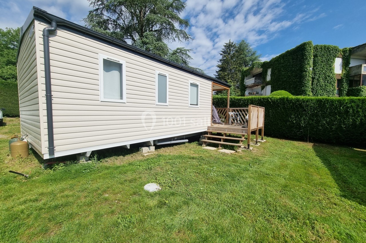 Mobil-home beige avec terrasse en bois, situé sur une pelouse entourée de haies et d'arbres sous un ciel partiellement…
