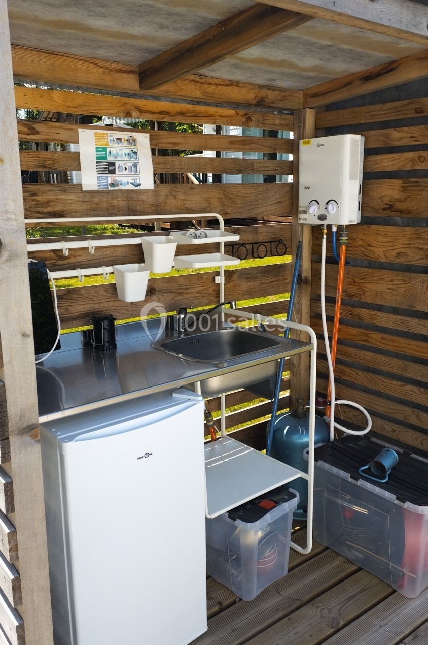 Cuisine extérieure en bois avec évier, réfrigérateur, chauffe-eau mural et étagères de rangement.