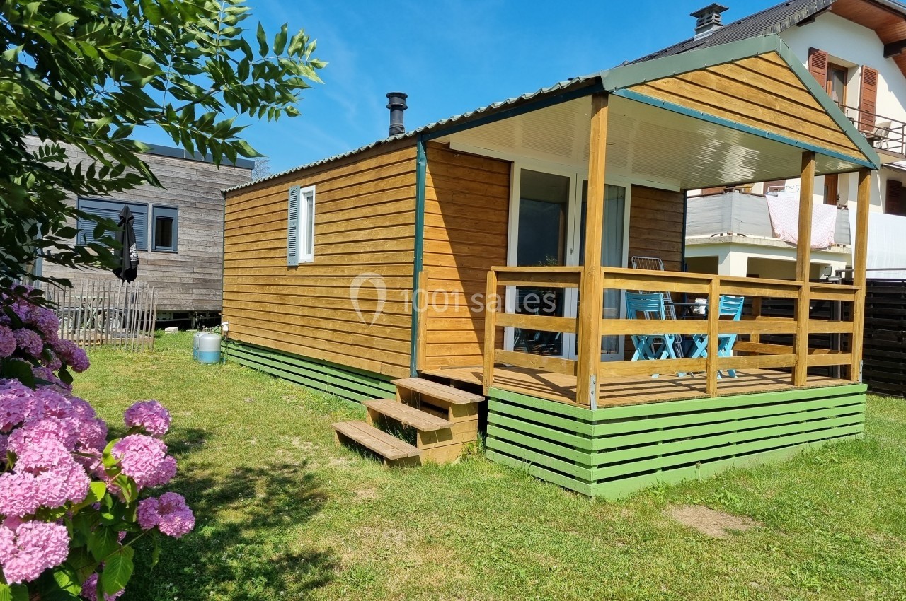 Petit chalet en bois avec terrasse couverte, entouré de pelouse et de fleurs, situé près d'autres habitations.