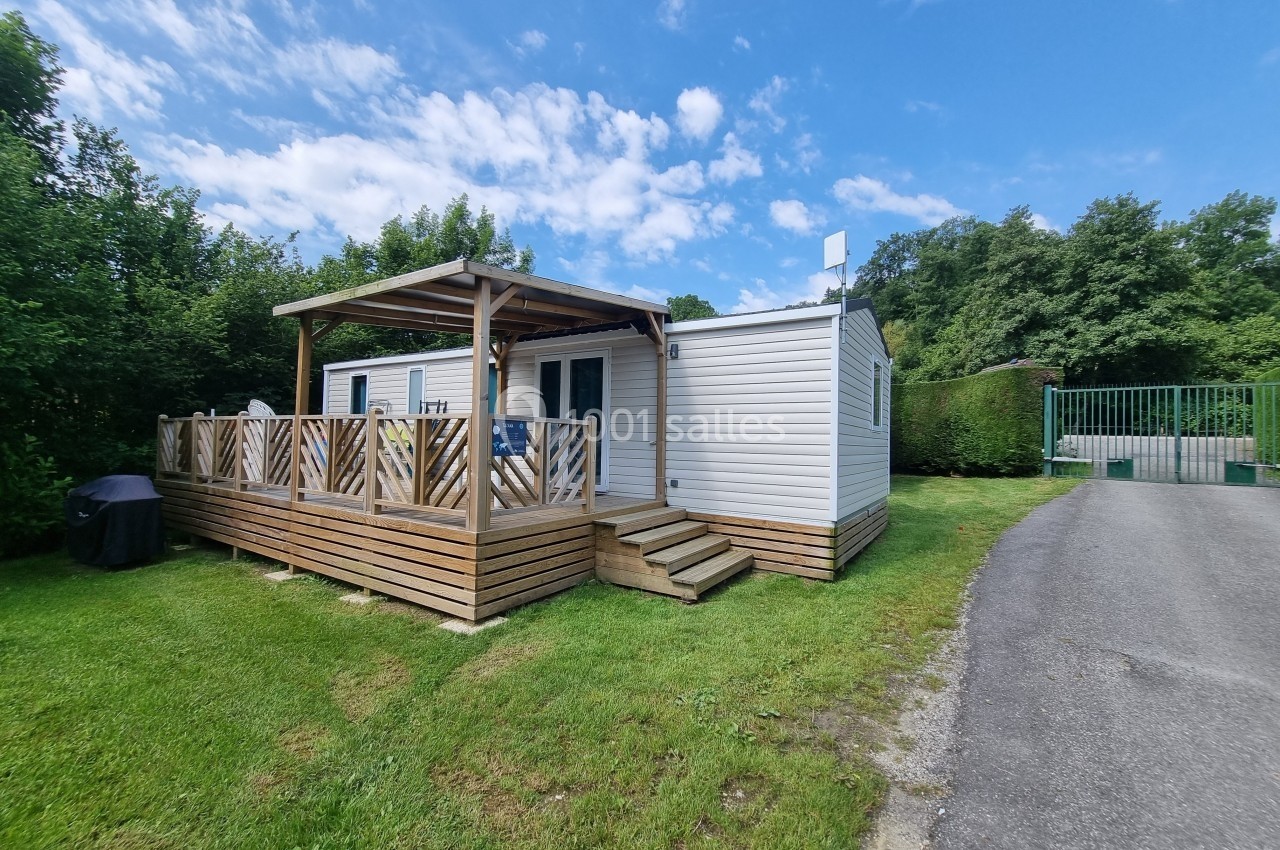 Mobil-home blanc avec terrasse en bois, situé sur une pelouse près d'une route et entouré de verdure.