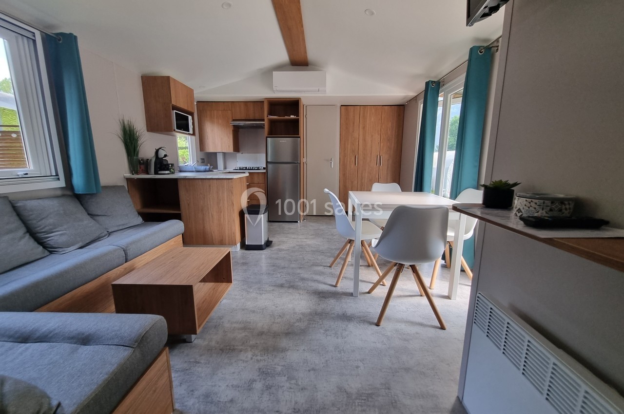 Intérieur d'un mobil-home moderne avec salon, cuisine équipée, table à manger et grandes fenêtres laissant entrer la lumière.