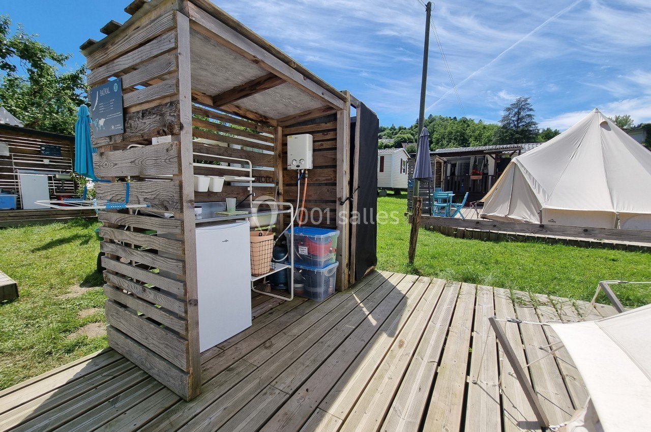 Cuisine extérieure en bois avec évier, réfrigérateur et étagères, située sur une terrasse en bois dans un camping.