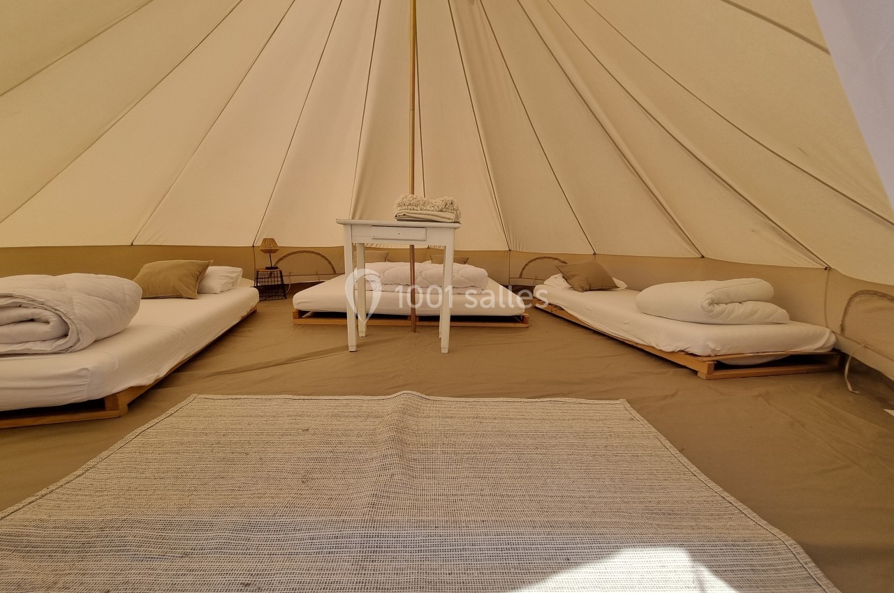 Intérieur d'une tente spacieuse avec lits simples, table centrale, tapis clair et lumière naturelle.