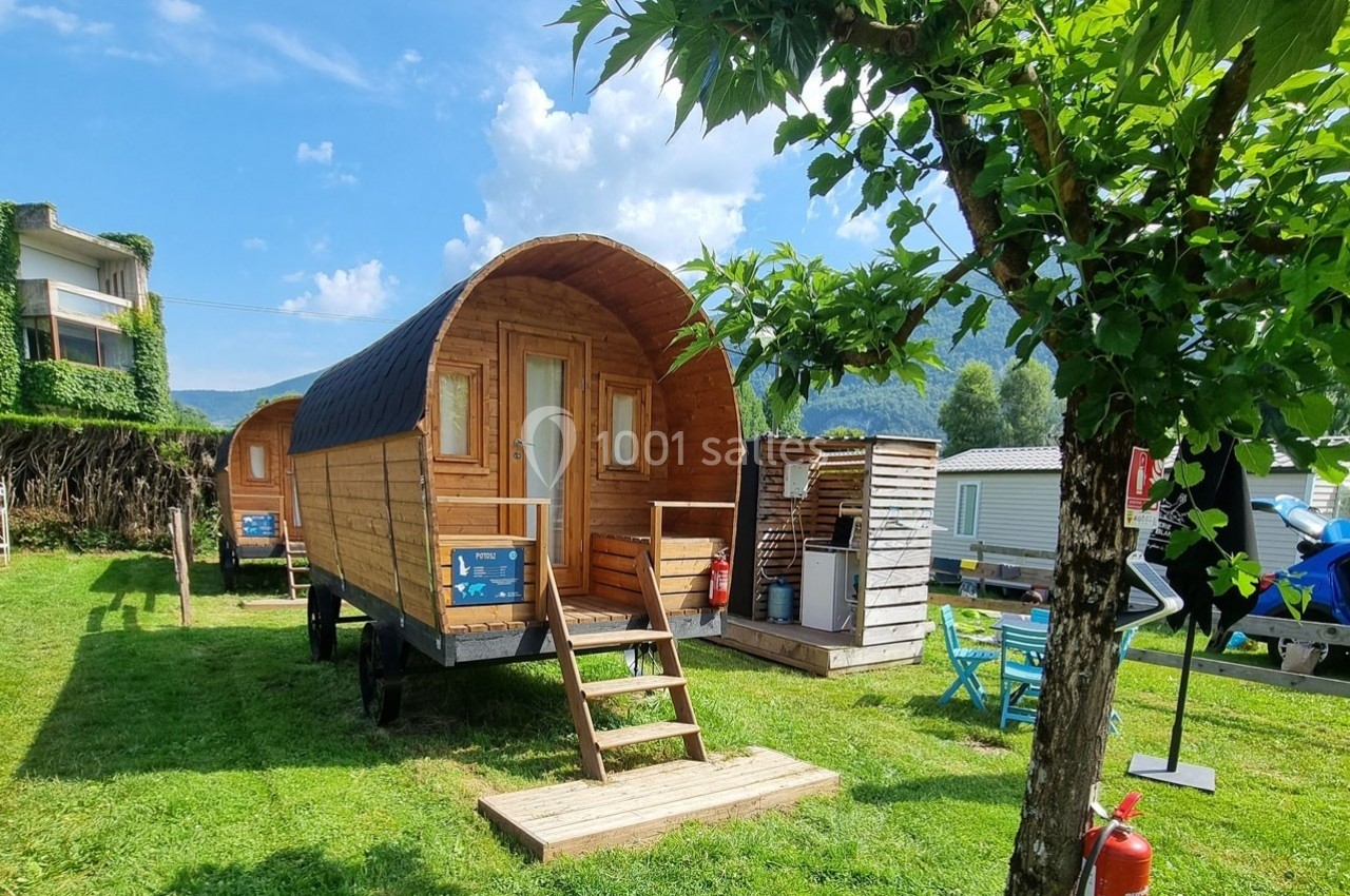 Roulotte en bois installée sur une pelouse avec terrasse, entourée d'arbres et de petites structures en bois.