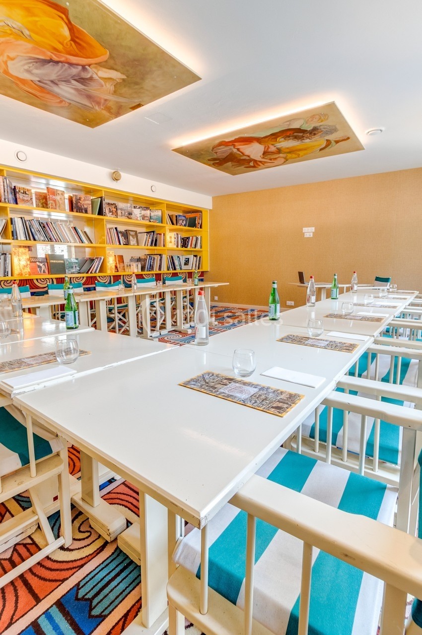 Salle de réunion lumineuse avec tables blanches, chaises colorées, étagères remplies de livres et fresques au plafond.