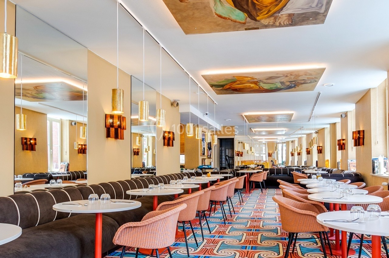 Salle de restaurant lumineuse avec tables rondes, chaises colorées, fresques au plafond et sol à motifs géométriques.