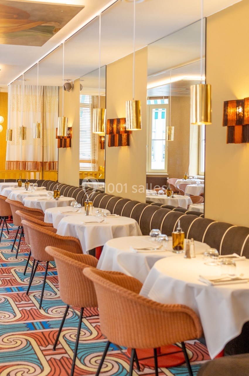 Salle de restaurant élégante avec tables dressées, chaises en tissu, miroirs muraux et éclairage chaleureux.