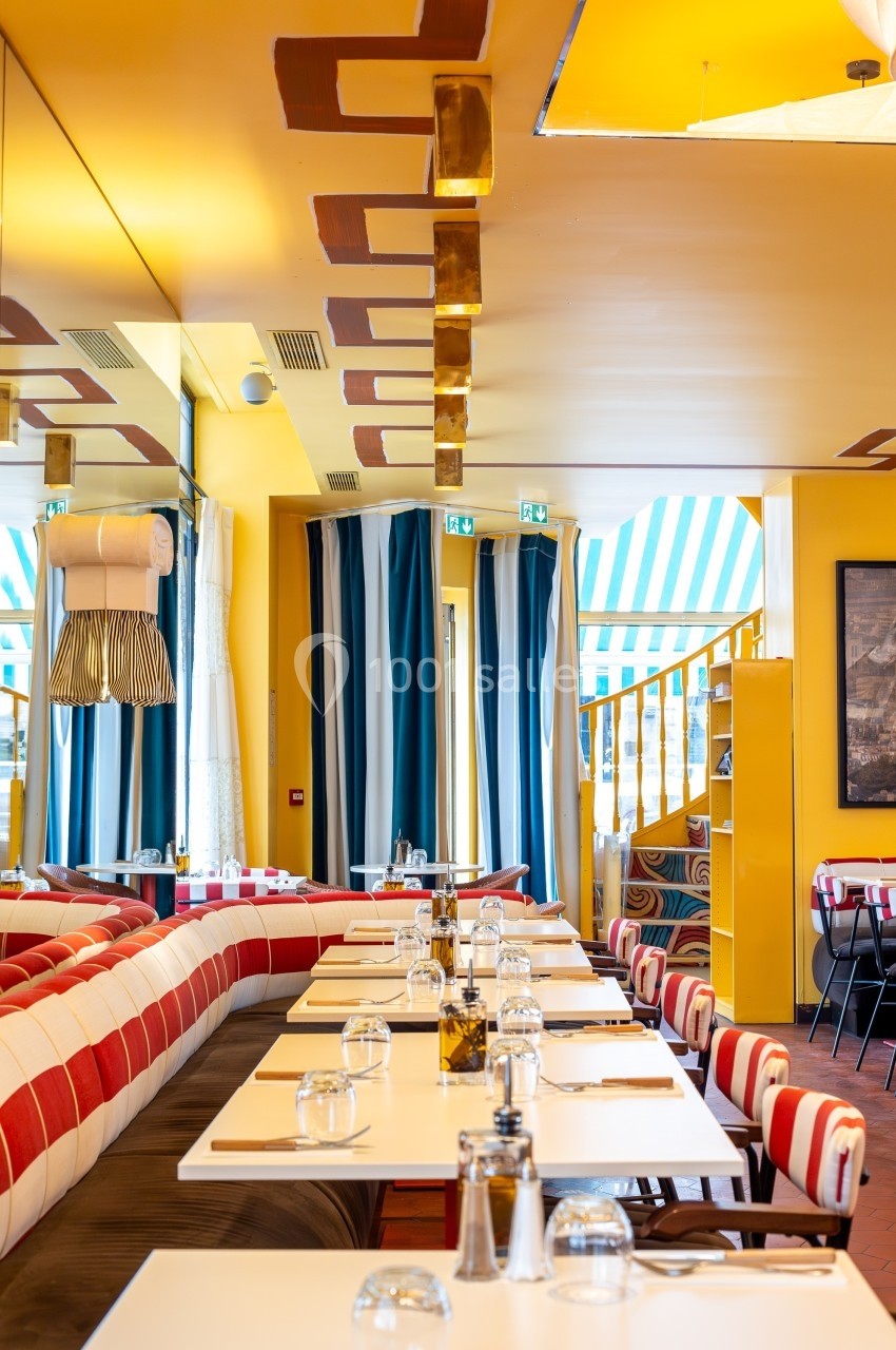 Salle de restaurant lumineuse avec tables dressées, banquettes rayées rouges et blanches, et décor coloré.