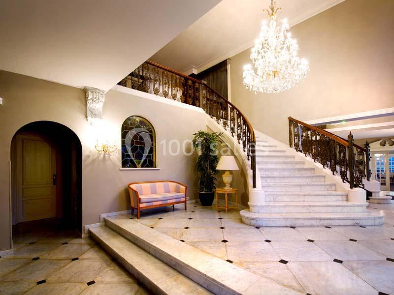 Escalier en marbre avec rampe en fer forgé, lustre imposant, canapé rayé et plante dans un hall élégant.