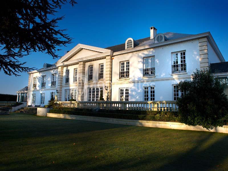 Façade d'un grand bâtiment blanc de style classique entouré d'un jardin verdoyant sous un ciel bleu.