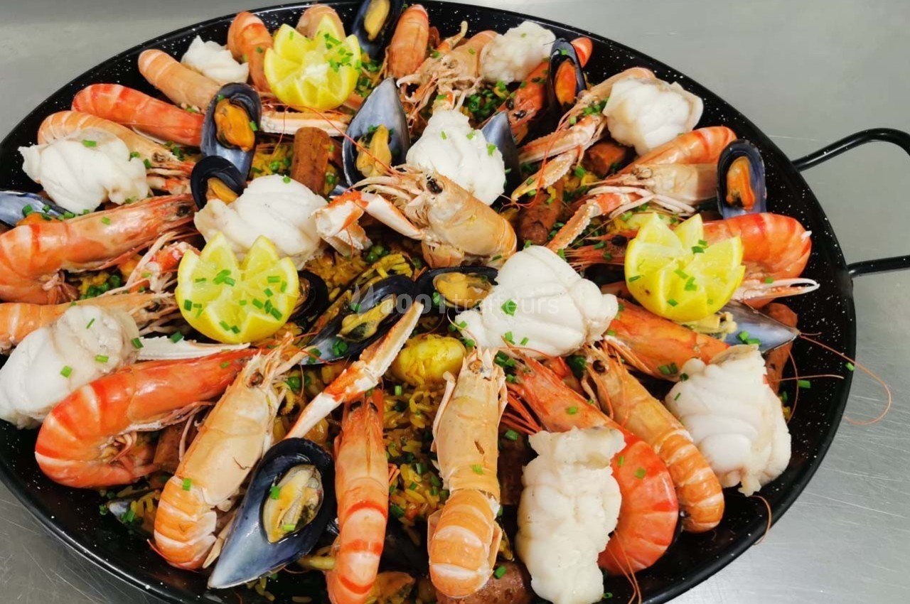 Plat de paella garni de crevettes, moules, morceaux de poisson et quartiers de citron, présenté dans une poêle.