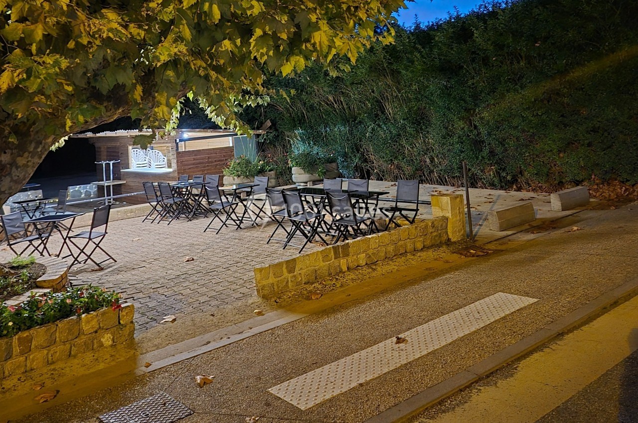 Terrasse extérieure avec tables et chaises sous un arbre, éclairée le soir, bordée de végétation et d'un chemin.