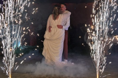 Un couple en tenue de mariage danse dans une salle éclairée par des lumières colorées et entourée de fumée.