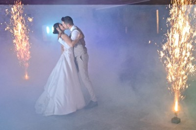 Un couple en tenue de mariage danse dans une salle éclairée par des lumières colorées et entourée de fumée.