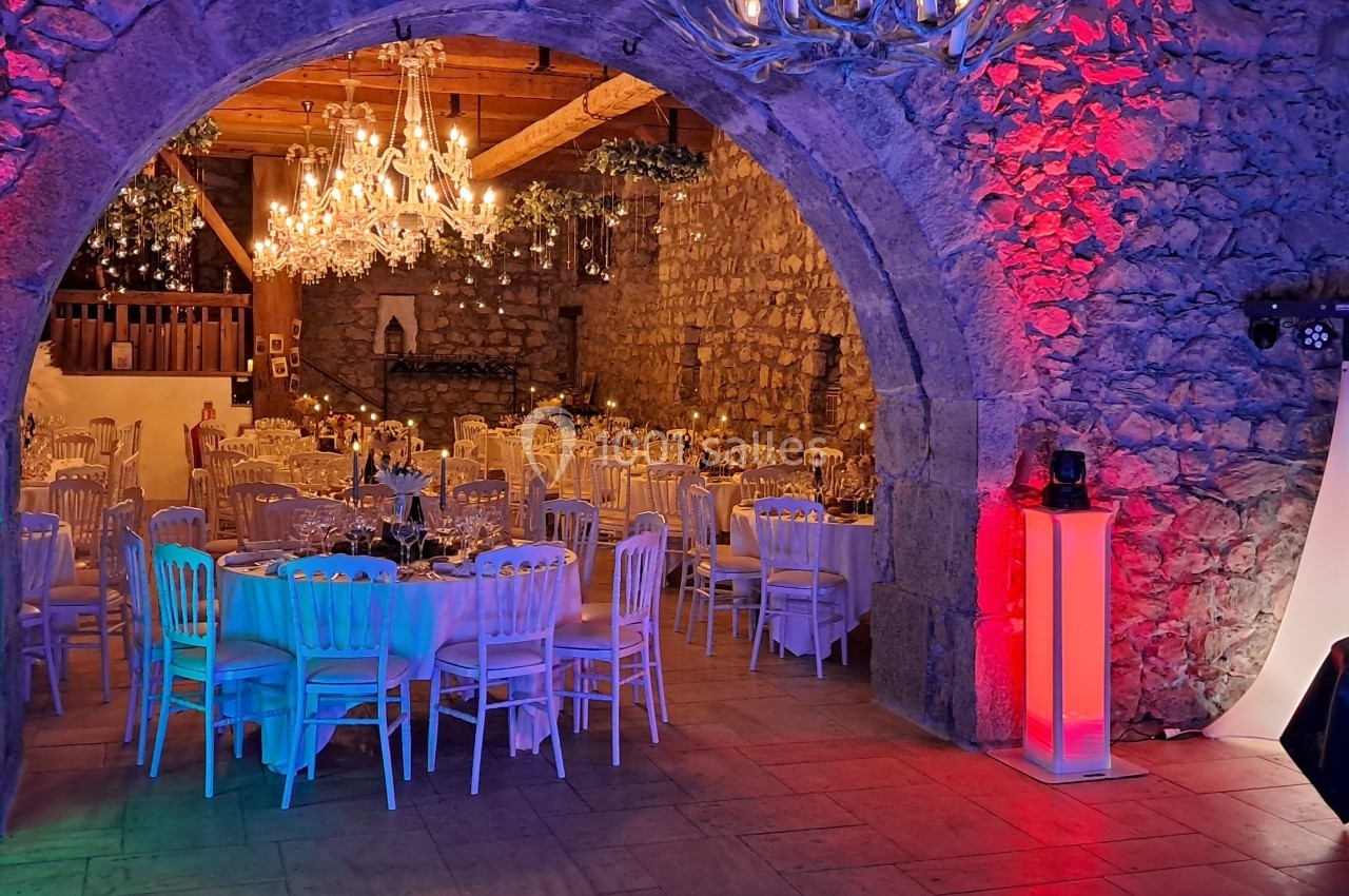 Salle en pierre décorée pour un événement, avec tables dressées, chandeliers et éclairages colorés rouge et bleu.