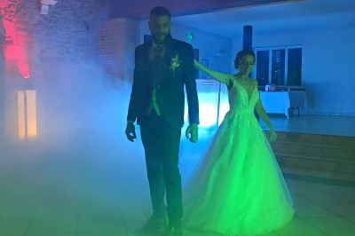 Un couple en tenue de mariage danse dans une salle éclairée par des lumières colorées et entourée de fumée.