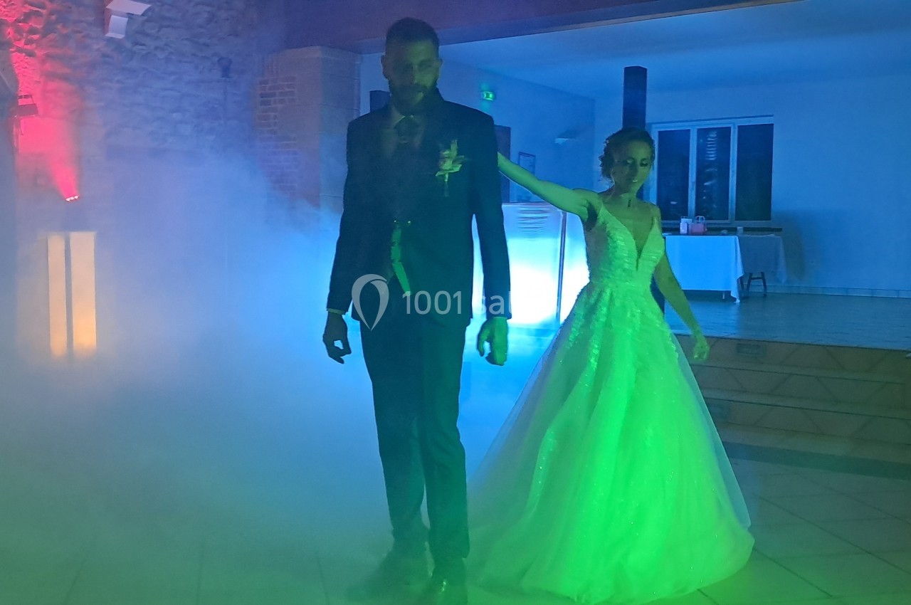 Un couple en tenue de mariage danse dans une salle éclairée par des lumières colorées et entourée de fumée.