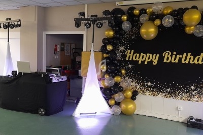 Décoration d'anniversaire avec ballons noirs, dorés et blancs, panneau ’Happy Birthday’ et matériel de sonorisation.