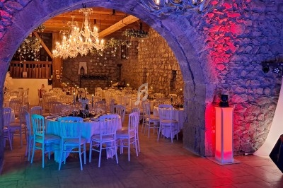 Salle en pierre éclairée par des lumières colorées, avec des tables rondes décorées pour un événement festif.
