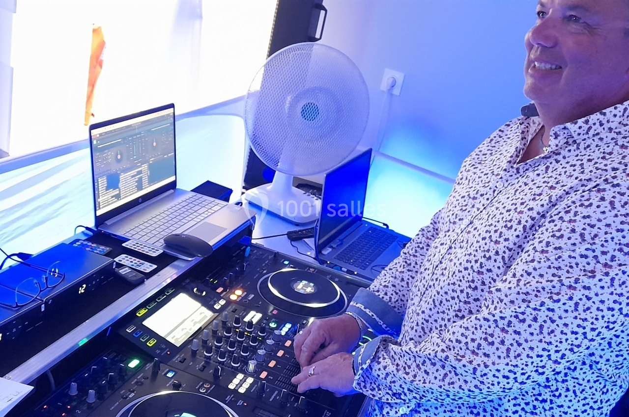 Un DJ souriant mixe de la musique sur une console avec des ordinateurs dans un environnement lumineux.