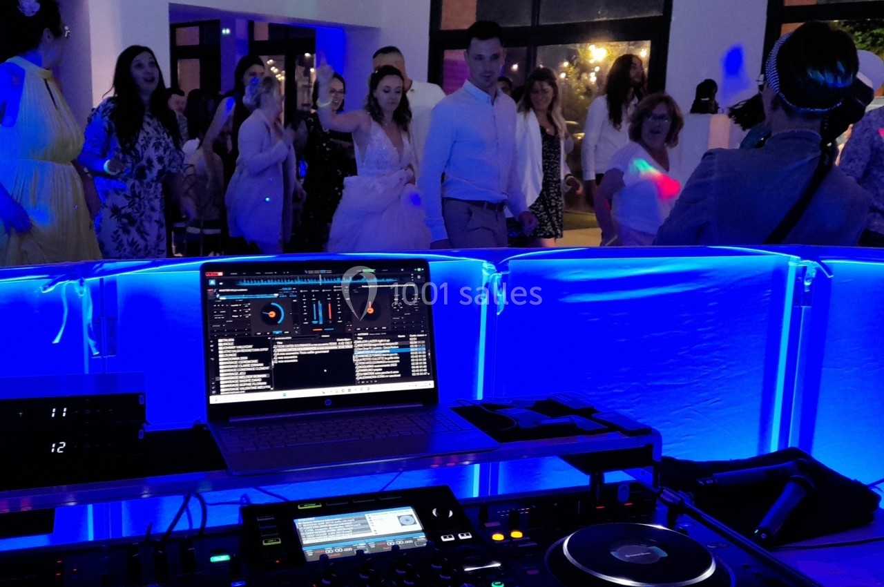 Vue depuis la table du DJ avec un ordinateur et des équipements audio, des invités dansant en arrière-plan sous des lumières…