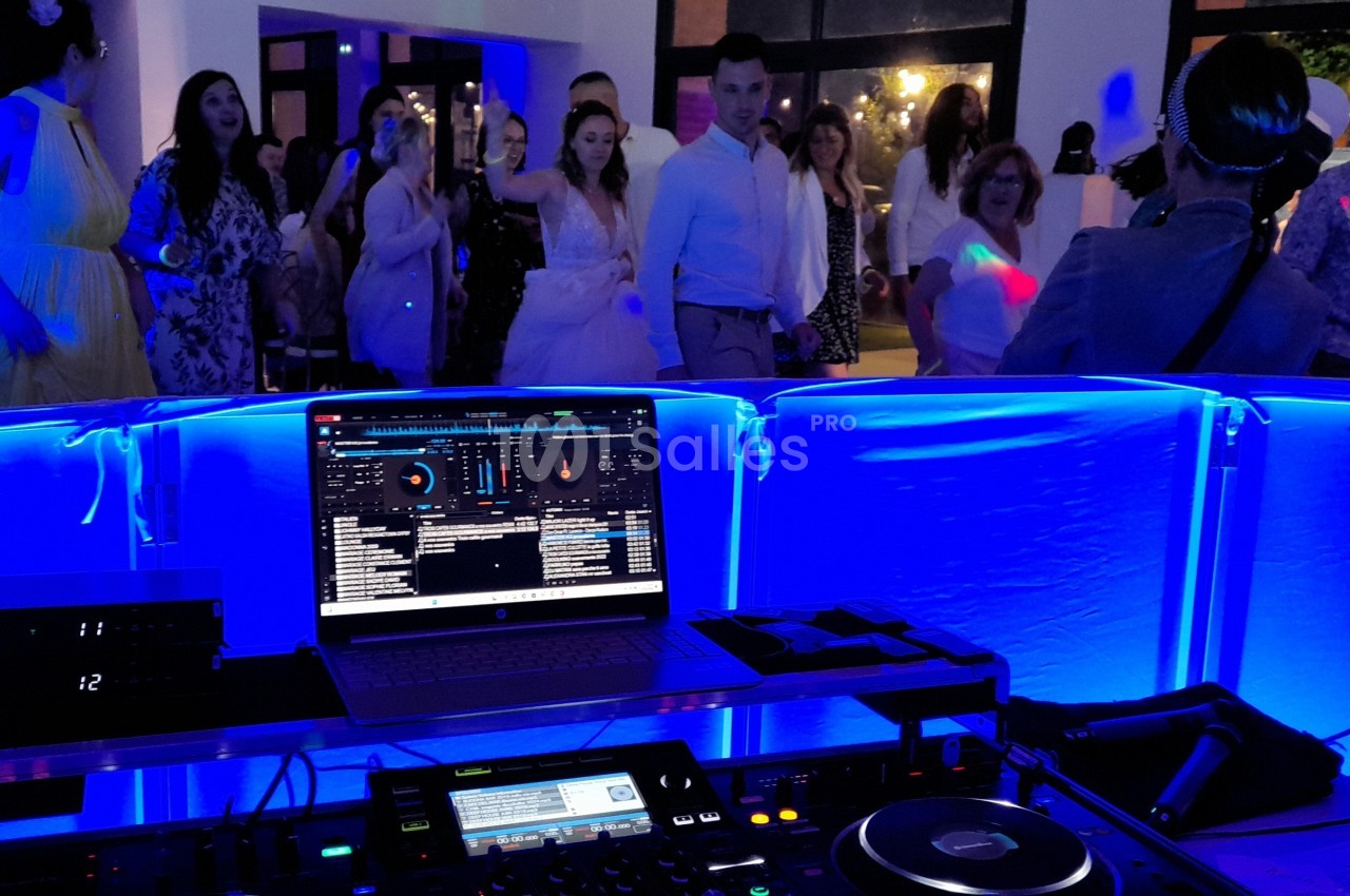 Vue depuis la table du DJ avec un ordinateur et des équipements audio, des invités dansant en arrière-plan sous des lumières…