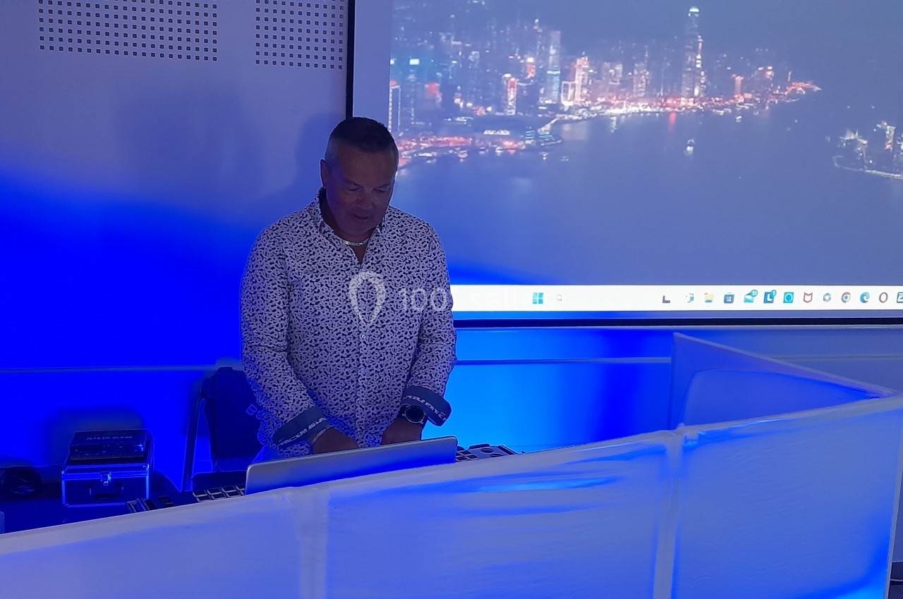 Homme debout devant un ordinateur portable, dans une salle éclairée par des lumières bleues, avec une projection en arrière…