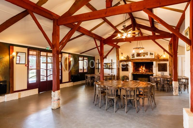 Salle spacieuse avec charpente en bois apparente, tables et chaises en bois, cheminée allumée en arrière-plan.