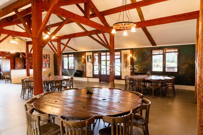 Salle lumineuse avec poutres apparentes, grandes tables en bois rondes et chaises, décor rustique et fenêtres donnant sur l…