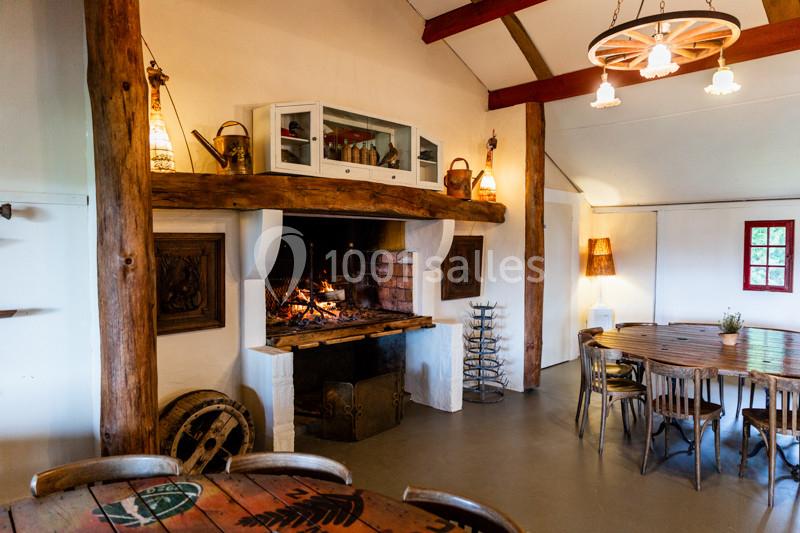 Salle rustique avec cheminée en pierre, poutres apparentes, tables en bois et éclairage chaleureux.