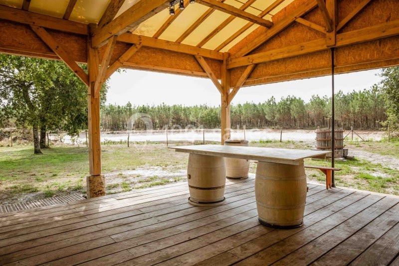 Terrasse en bois couverte avec une table faite de tonneaux, offrant une vue sur un étang et une forêt en arrière-plan.