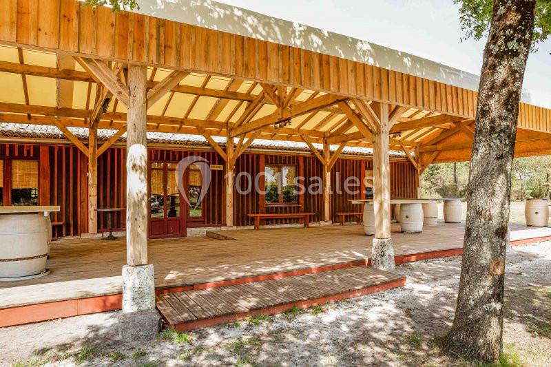 Terrasse en bois couverte avec structure en poutres, entourée d'arbres, devant un bâtiment en bois.