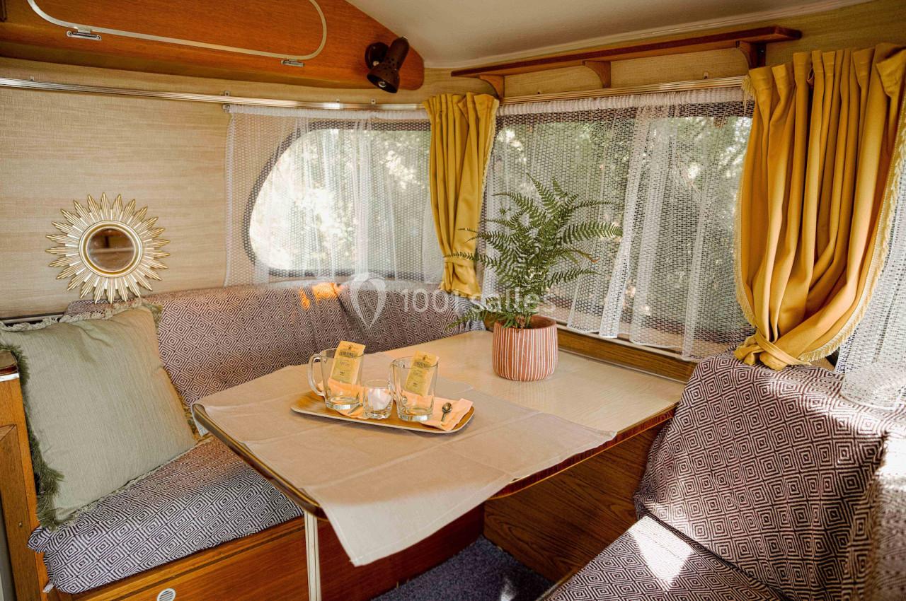 Intérieur d'une caravane avec banquettes, table dressée, plante en pot et rideaux jaunes laissant passer la lumière.