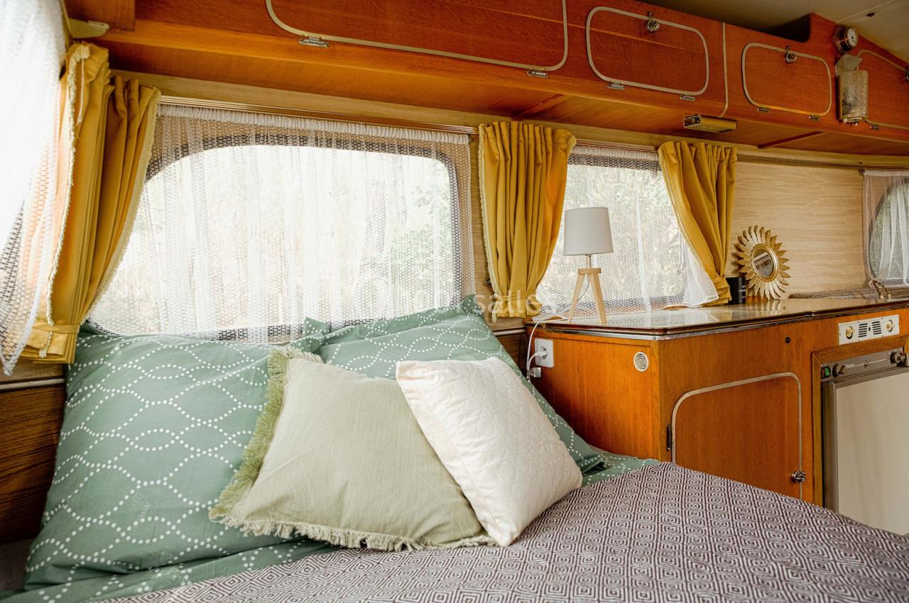 Intérieur d'une caravane avec lit, coussins verts, rideaux jaunes, lampe de chevet et mobilier en bois.