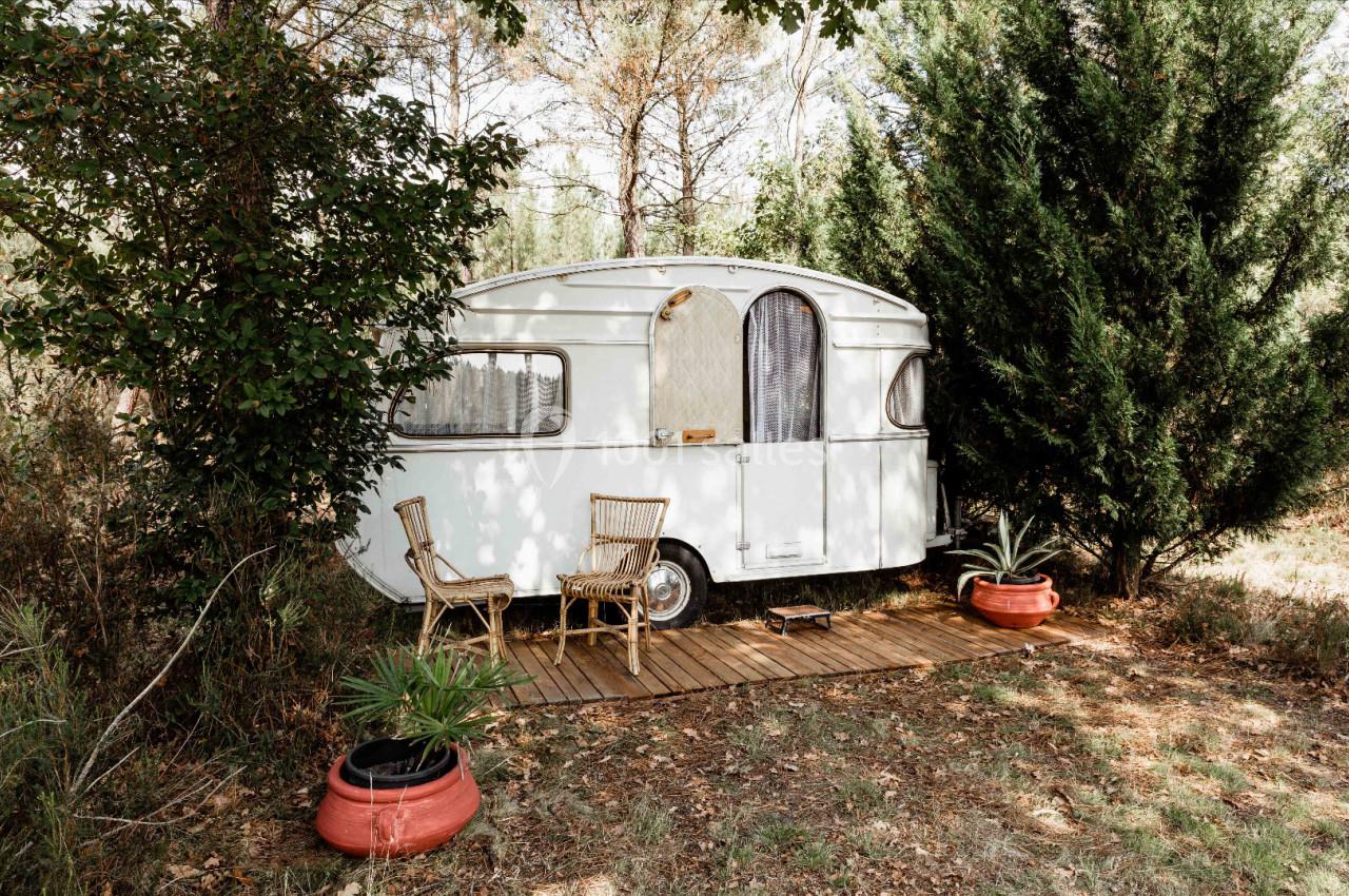 Caravane blanche stationnée dans un environnement boisé, avec deux chaises en rotin sur une terrasse en bois.