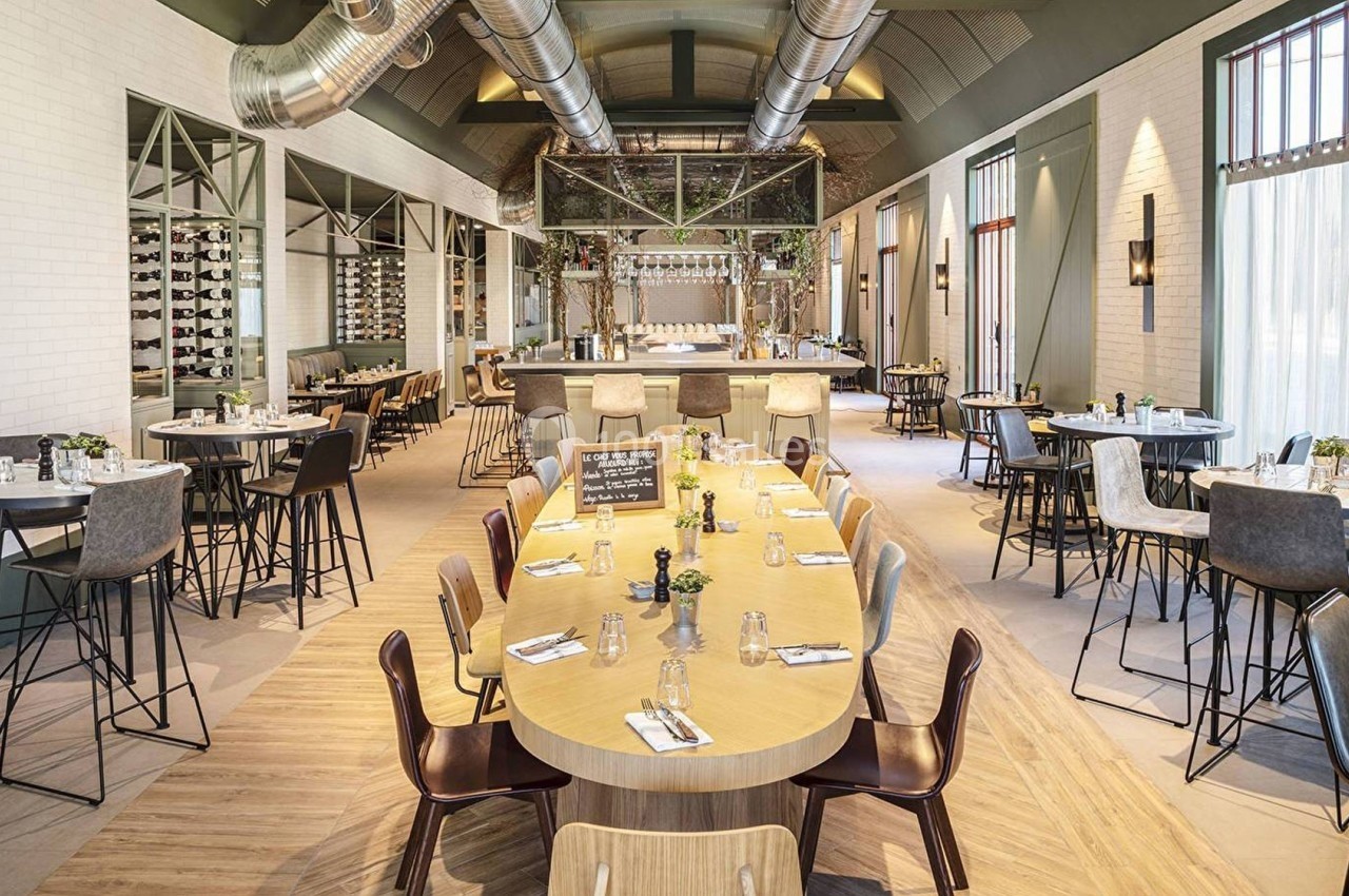 Salle de restaurant lumineuse avec tables en bois, chaises variées et bar central entouré de décorations végétales.