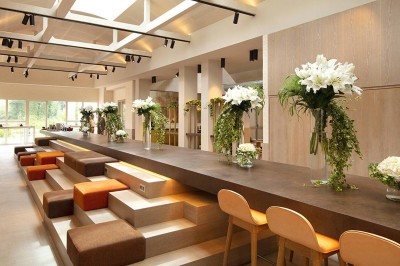 Salon lumineux avec canapé beige, table en bois, suspension noire, bouquet de fleurs et grandes fenêtres.