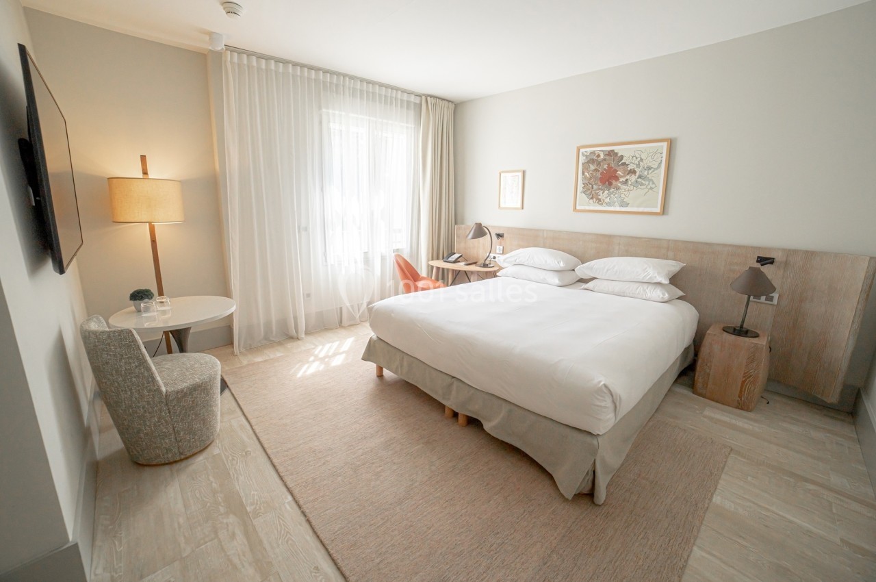 Chambre lumineuse avec un lit double, fauteuil, bureau, lampe et grande fenêtre avec rideaux translucides.