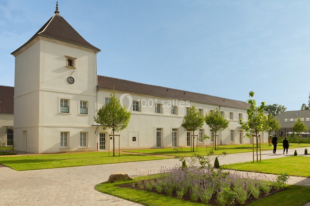 Bâtiment blanc à deux étages avec une tour, entouré d'espaces verts et de chemins pavés, sous un ciel dégagé.