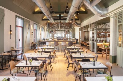 Salon lumineux avec canapé beige, table en bois, suspension noire, bouquet de fleurs et grandes fenêtres.