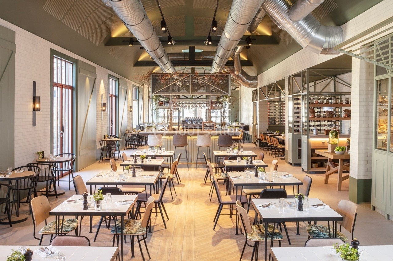 Salle de restaurant lumineuse avec tables dressées, chaises variées, plafond industriel et cuisine ouverte en arrière-plan.