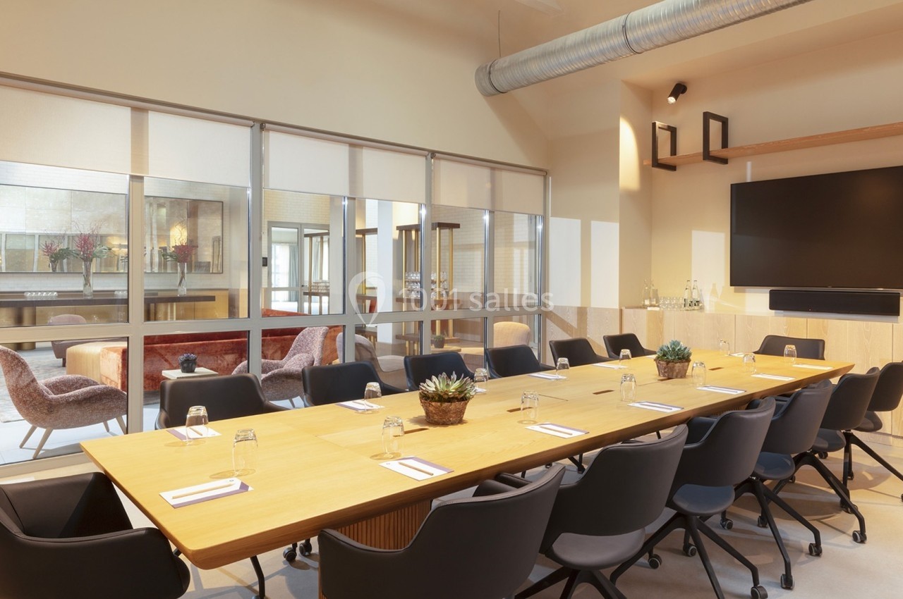 Salle de réunion moderne avec une grande table en bois, chaises noires, écran mural et cloison vitrée donnant sur un espace…
