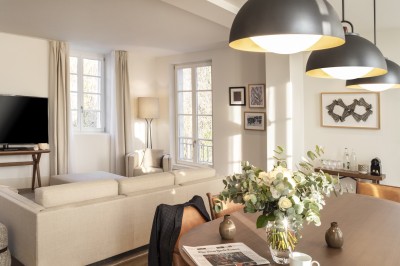 Salon lumineux avec canapé beige, table en bois, suspension noire, bouquet de fleurs et grandes fenêtres.