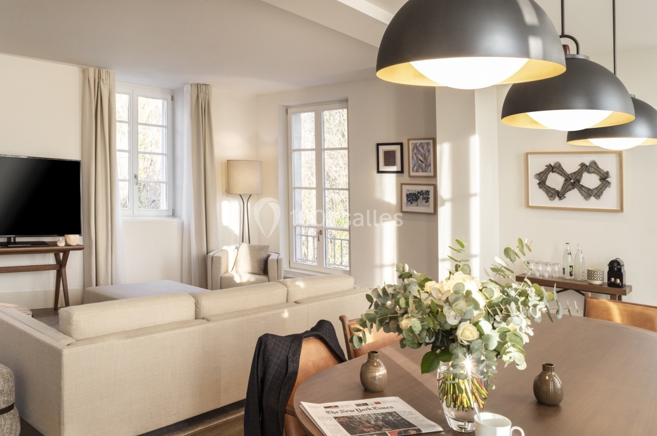 Salon lumineux avec canapé beige, table en bois, suspension noire, bouquet de fleurs et grandes fenêtres.
