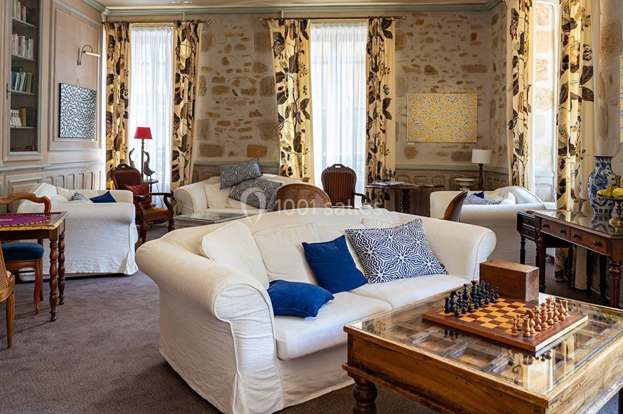 Salon lumineux avec canapés blancs, table d'échecs en bois, bibliothèque et murs en pierre apparente.